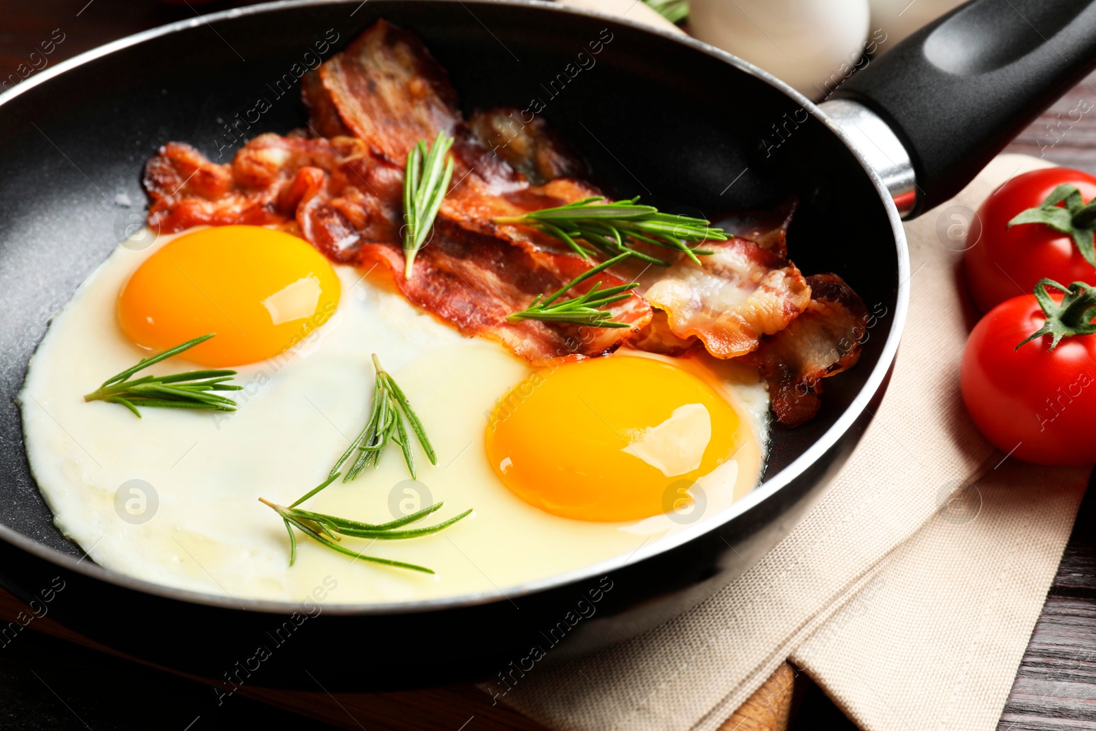 Photo of Tasty bacon, eggs and rosemary in frying pan near tomatoes on wooden table, closeup