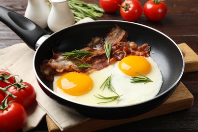 Photo of Tasty bacon, eggs and rosemary in frying pan near tomatoes on wooden table, closeup