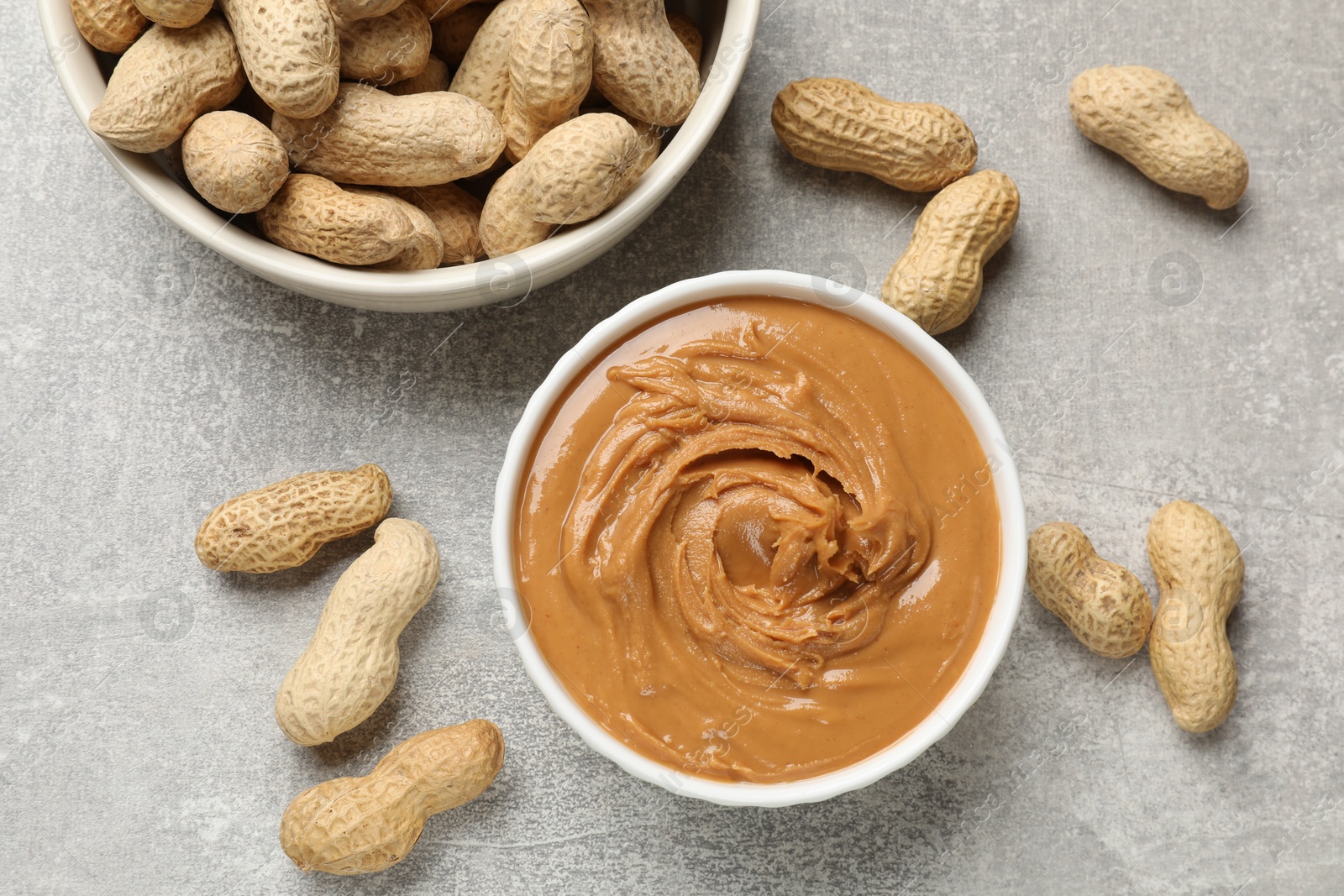 Photo of Tasty peanut butter in bowl and groundnuts on grey textured table, flat lay