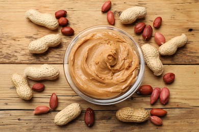 Tasty peanut butter in bowl and groundnuts on wooden table, flat lay