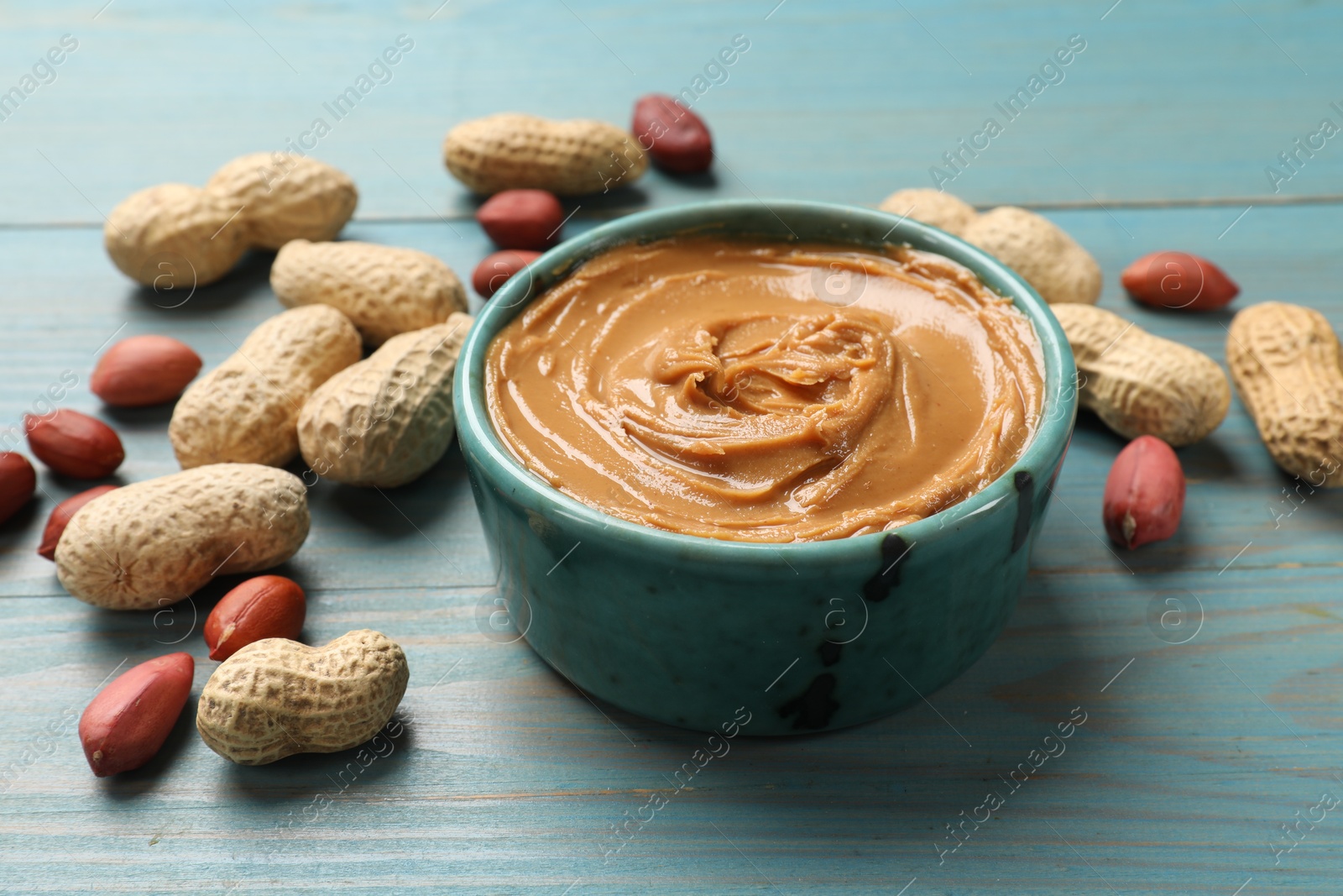 Photo of Tasty peanut butter in bowl and groundnuts on light blue wooden table, closeup