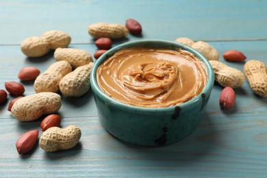 Tasty peanut butter in bowl and groundnuts on light blue wooden table, closeup