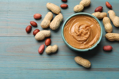 Tasty peanut butter in bowl and groundnuts on light blue wooden table, flat lay