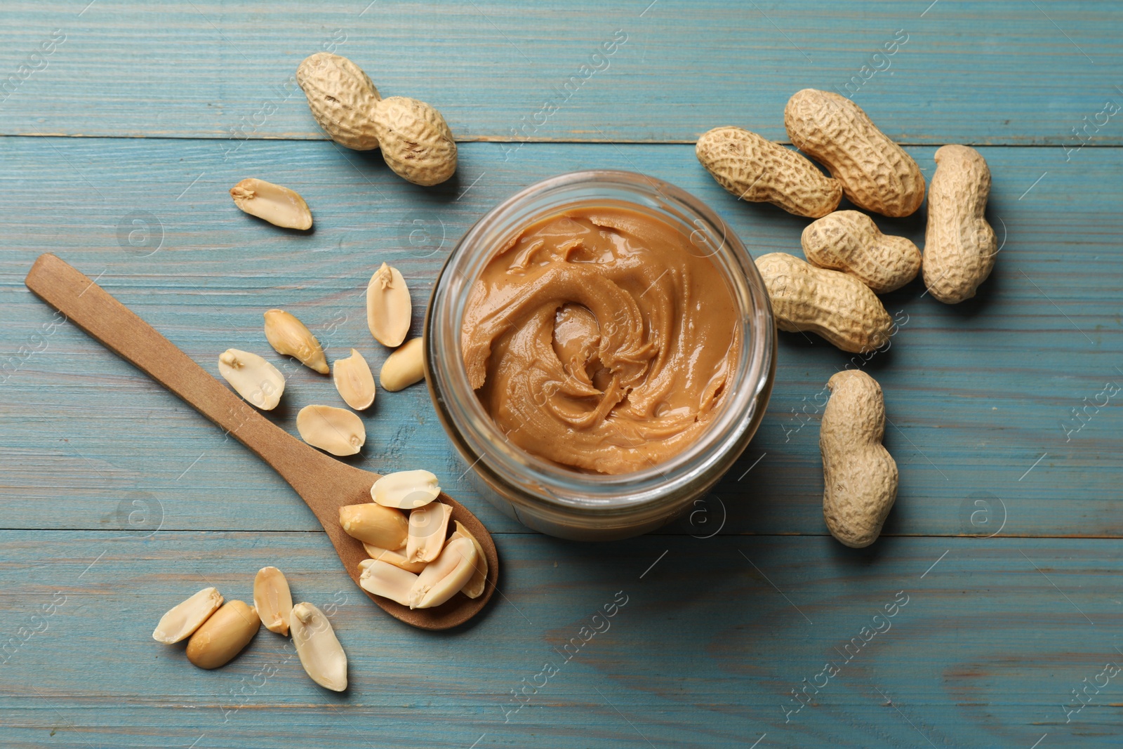 Photo of Tasty peanut butter in jar and groundnuts on blue wooden table, flat lay