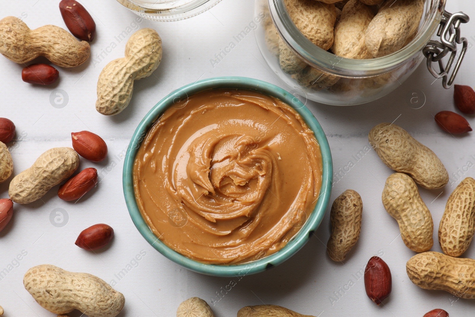 Photo of Tasty peanut butter in bowl and groundnuts on white tiled table, flat lay