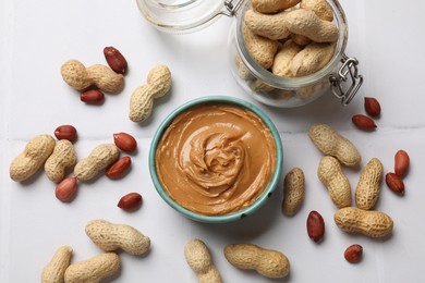 Tasty peanut butter in bowl and groundnuts on white tiled table, flat lay