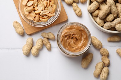 Tasty peanut butter in jar and groundnuts on white tiled table, flat lay