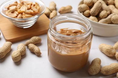 Tasty peanut butter in jar and groundnuts on white table