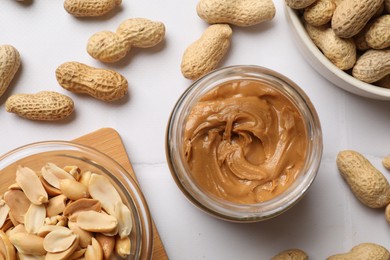 Tasty peanut butter in jar and groundnuts on white tiled table, flat lay