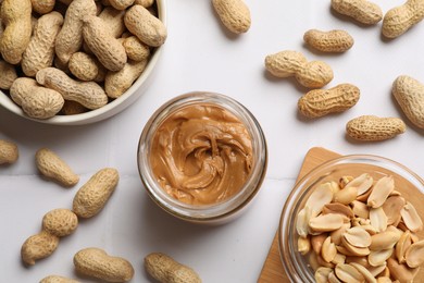 Tasty peanut butter in jar and groundnuts on white tiled table, flat lay