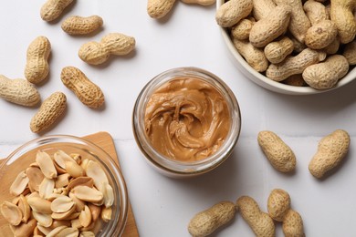 Photo of Tasty peanut butter in jar and groundnuts on white tiled table, flat lay