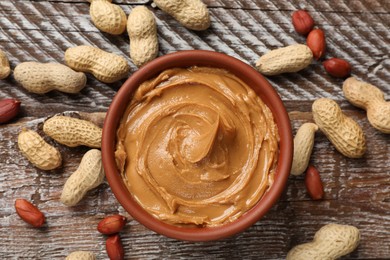 Tasty peanut butter in bowl and groundnuts on wooden table, flat lay