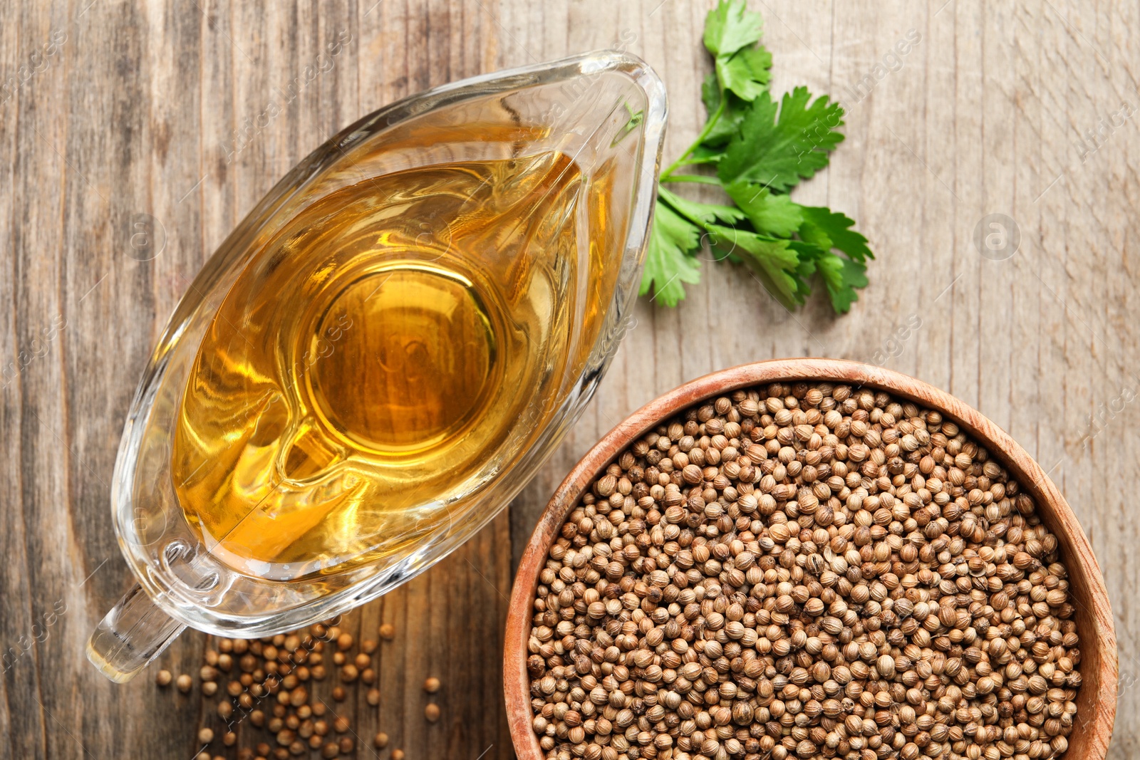 Photo of Dried coriander seeds in bowl, oil and green leaves on wooden table, top view