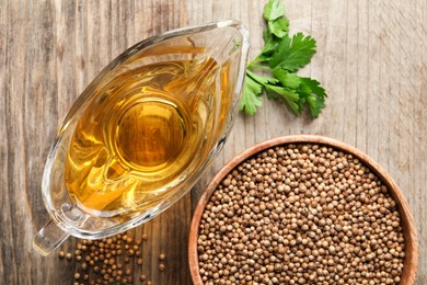 Dried coriander seeds in bowl, oil and green leaves on wooden table, top view