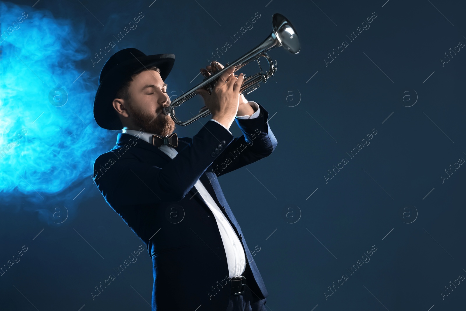 Photo of Professional musician playing trumpet on dark background in blue lights and smoke. Space for text