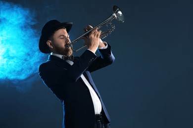 Photo of Professional musician playing trumpet on dark background in blue lights and smoke. Space for text