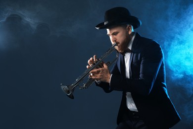 Photo of Professional musician playing trumpet on dark background in blue lights and smoke. Space for text
