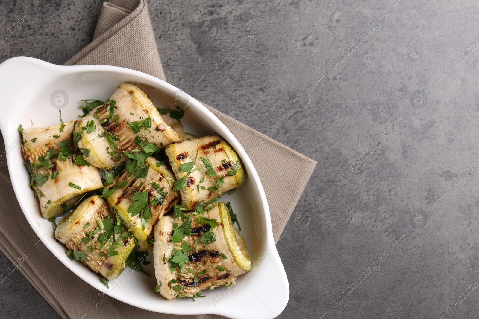 Photo of Tasty grilled zucchini rolls with parsley in dish on grey textured table, top view. Space for text