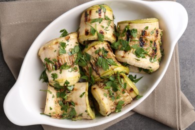 Tasty grilled zucchini rolls with parsley in dish on grey table, top view