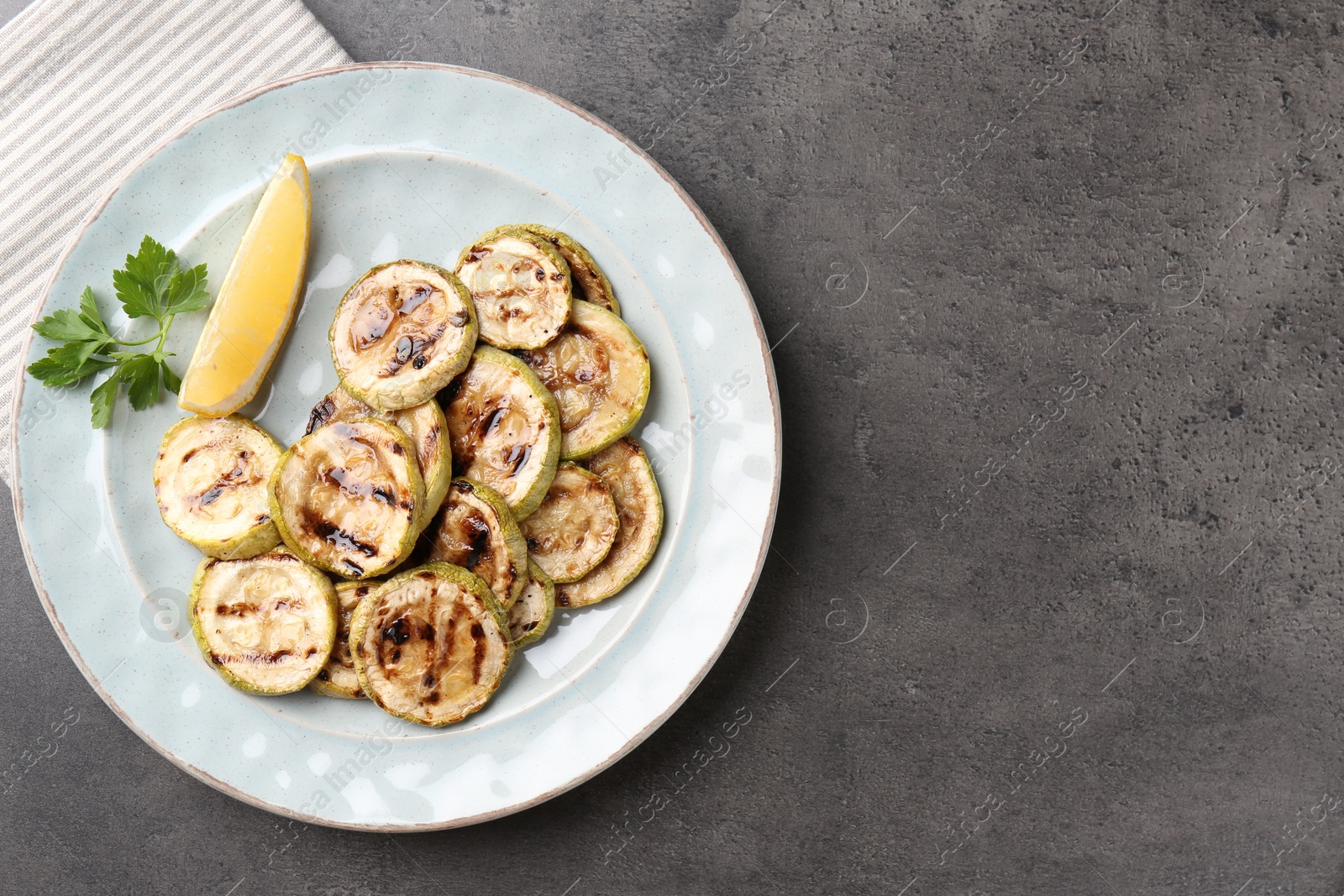 Photo of Tasty grilled zucchini slices with lemon and parsley on grey textured table, top view. Space for text