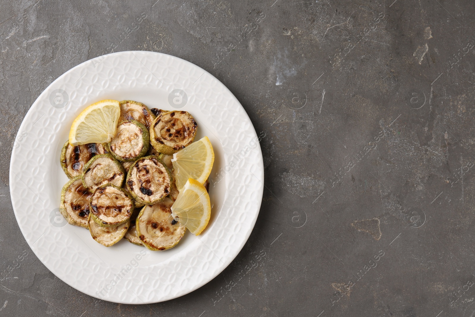 Photo of Tasty grilled courgette slices with lemon on grey textured table, top view. Space for text