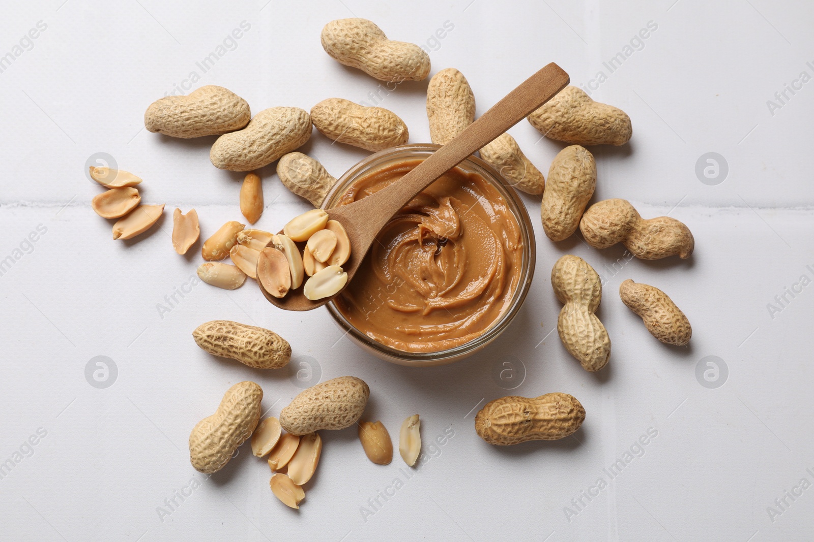 Photo of Tasty peanut butter in bowl and groundnuts on white tiled table, flat lay