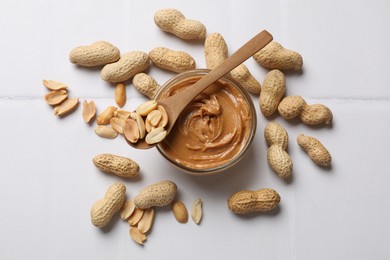 Photo of Tasty peanut butter in bowl and groundnuts on white tiled table, flat lay