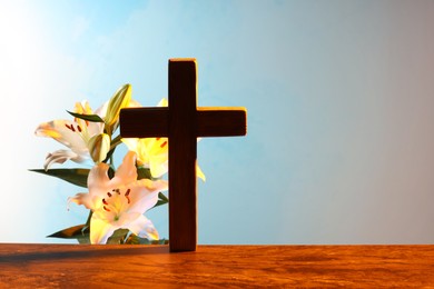 Wooden cross and beautiful lily flowers on table against light blue background, space for text. Religion of Christianity