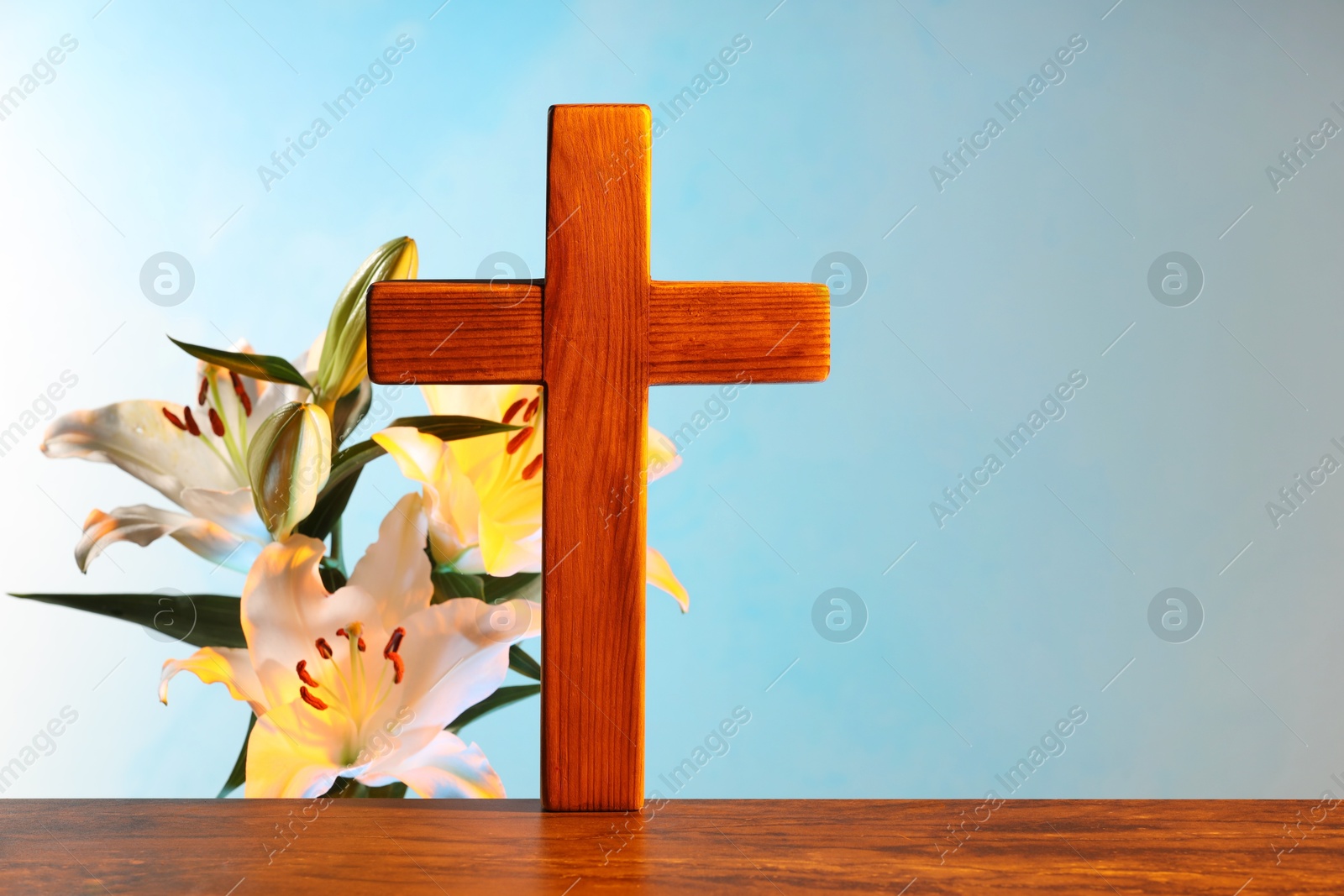 Photo of Wooden cross and beautiful lily flowers on table against light blue background, space for text. Religion of Christianity