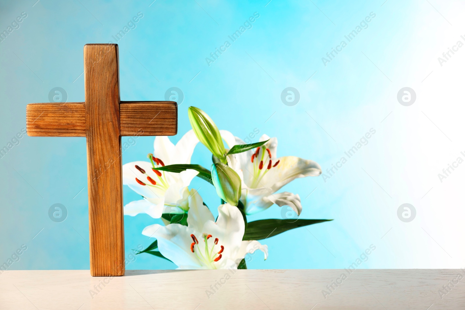 Photo of Wooden cross and beautiful lily flowers on table against light blue background, space for text. Religion of Christianity