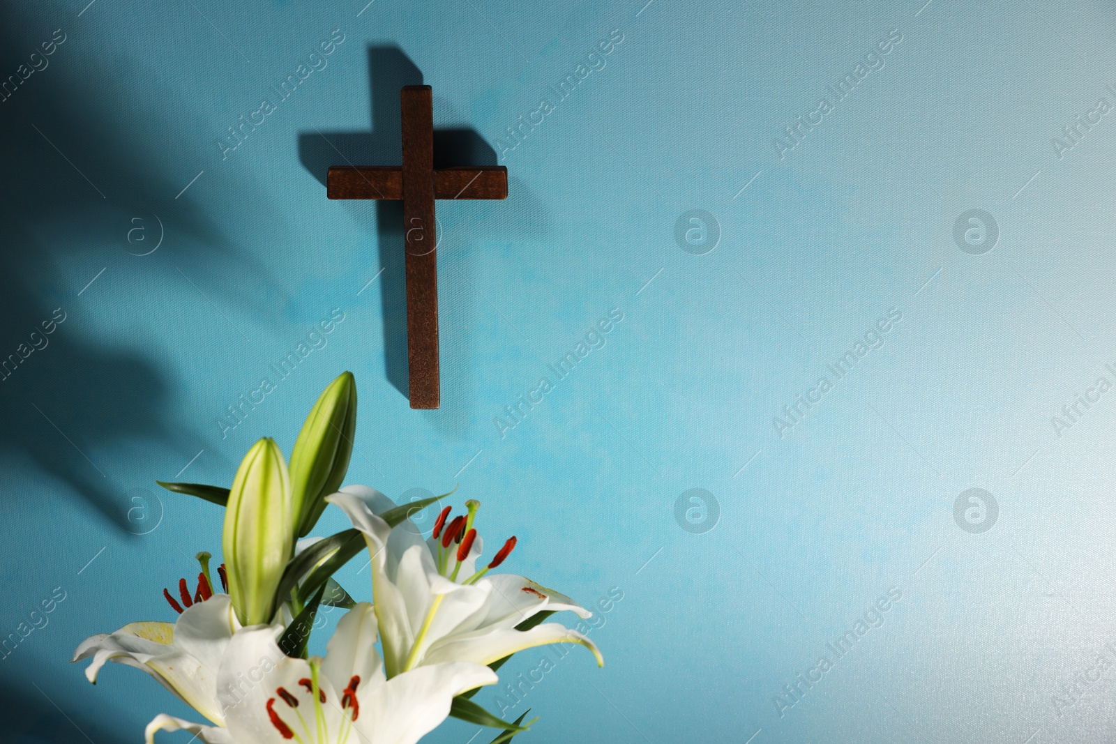 Photo of Wooden cross and lily flowers on light blue background, space for text. Religion of Christianity