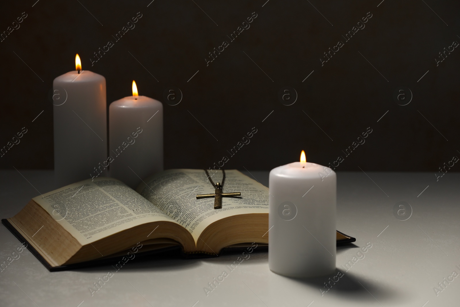 Photo of Cross with chain, burning candles and Bible on light table against black background. Religion of Christianity