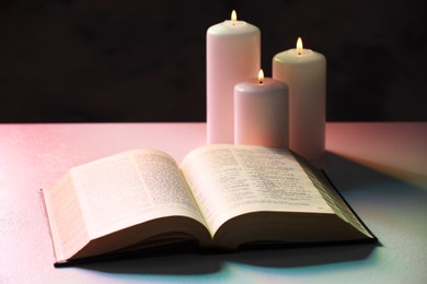 Photo of Bible and burning candles on light table against black background, closeup. Religion of Christianity