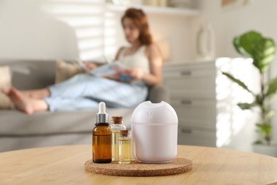 Photo of Woman reading magazine at home, focus on bottles of essential oils and aroma diffuser