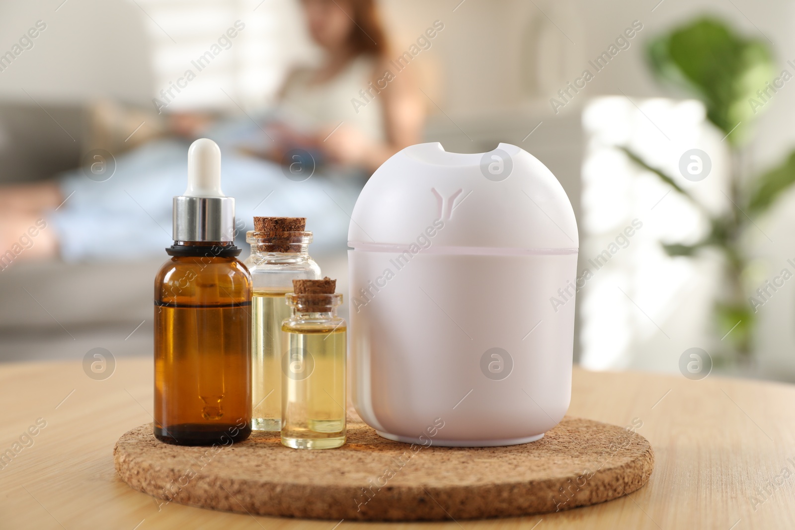 Photo of Woman reading magazine at home, focus on bottles of essential oils and aroma diffuser