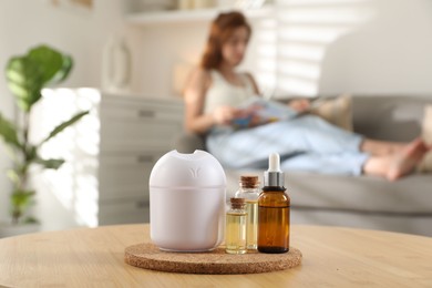 Photo of Woman reading magazine at home, focus on bottles of essential oils and aroma diffuser