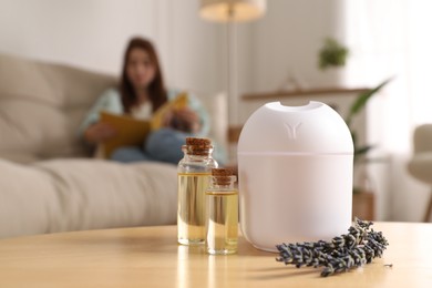 Woman reading magazine at home, focus on aroma diffuser, bottles of essential oils and lavender flowers