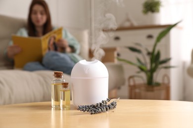 Woman reading magazine at home, focus on aroma diffuser, bottles of essential oils and lavender flowers