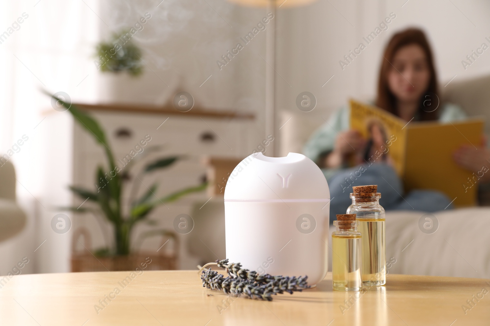 Photo of Woman reading magazine at home, focus on aroma diffuser, bottles of essential oils and lavender flowers