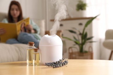 Woman reading magazine at home, focus on aroma diffuser, bottles of essential oils and lavender flowers