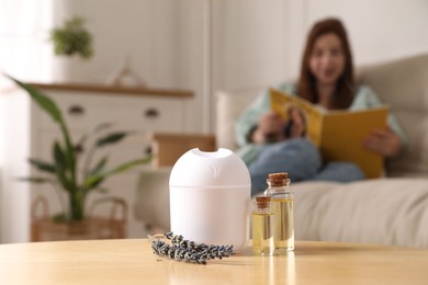 Woman reading magazine at home, focus on aroma diffuser, bottles of essential oils and lavender flowers