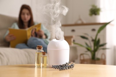 Woman reading magazine at home, focus on aroma diffuser, bottles of essential oils and lavender flowers