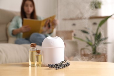 Woman reading magazine at home, focus on aroma diffuser, bottles of essential oils and lavender flowers