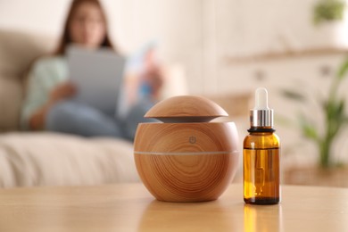 Photo of Woman reading magazine at home, focus on aroma diffuser and bottle of essential oil