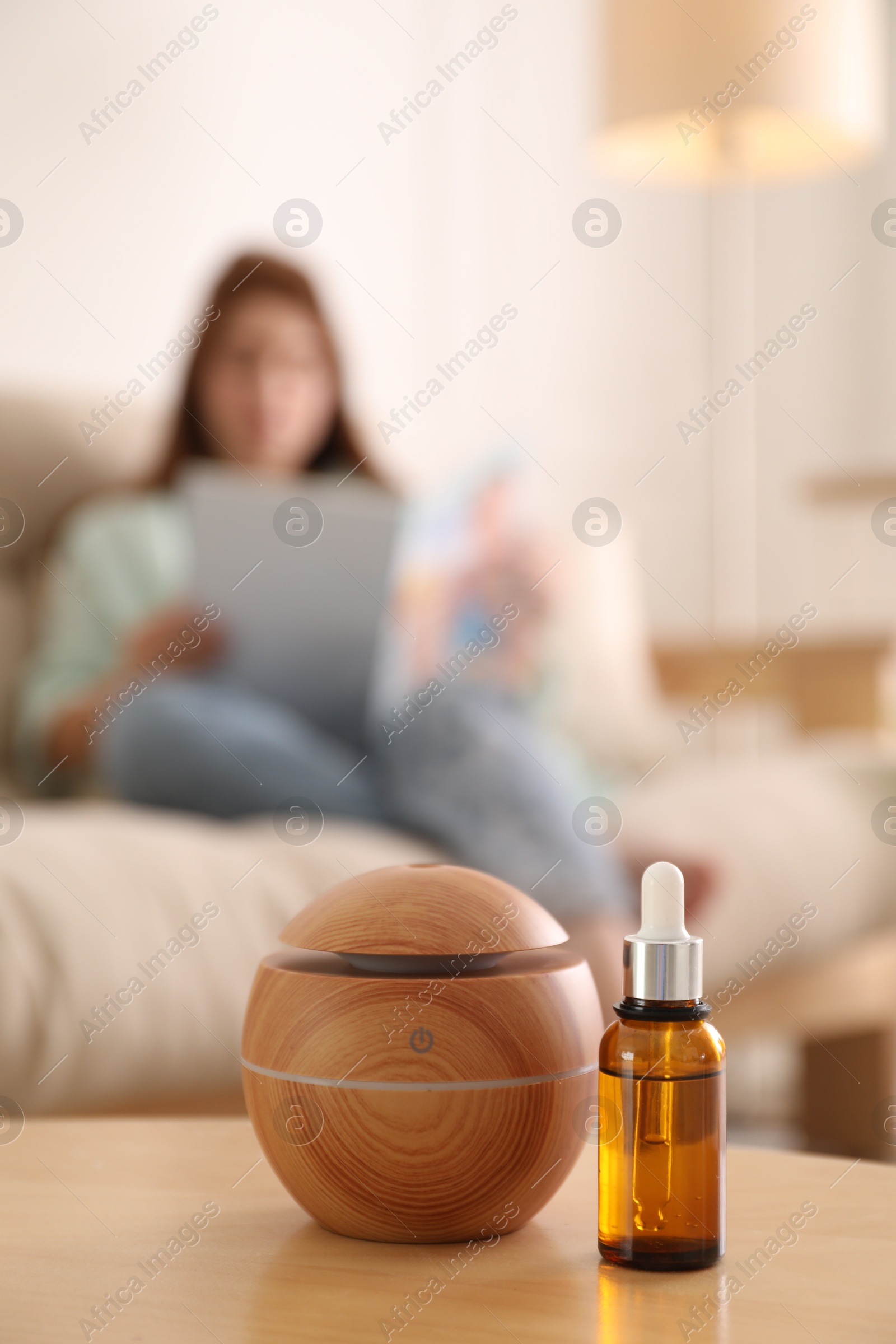 Photo of Woman reading magazine at home, focus on aroma diffuser and bottle of essential oil