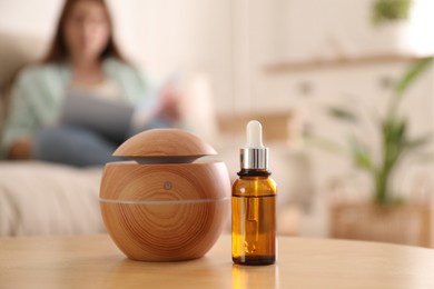 Photo of Woman reading magazine at home, focus on aroma diffuser and bottle of essential oil