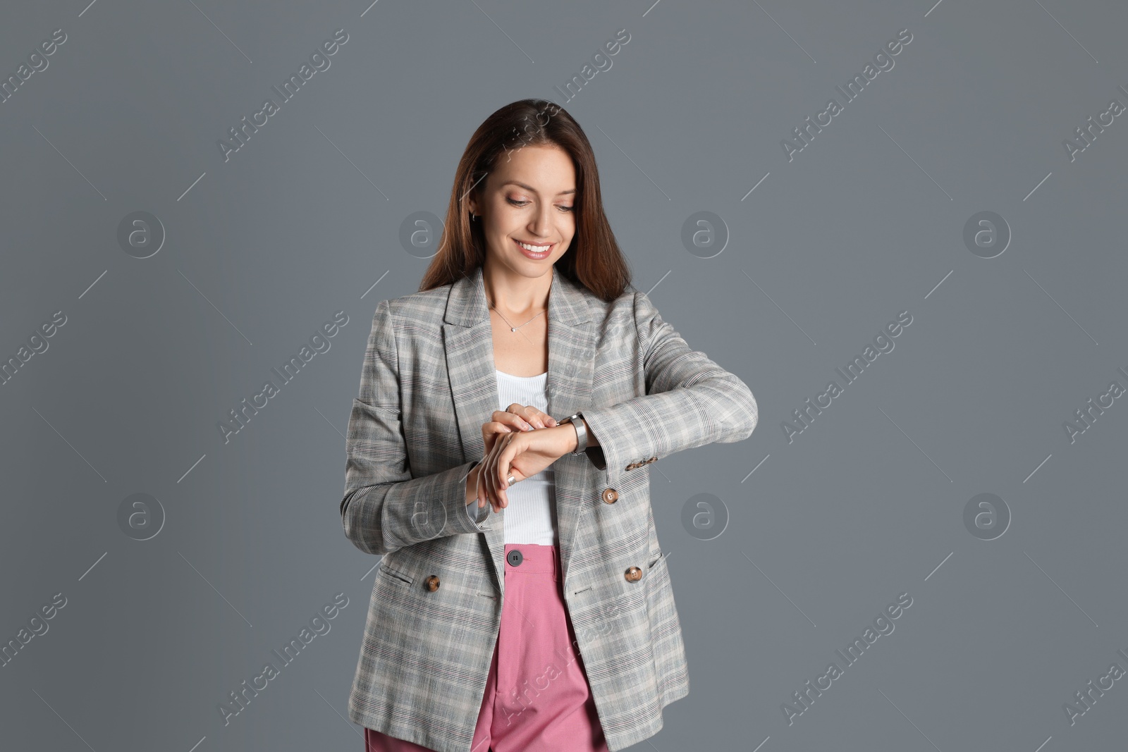 Photo of Beautiful woman in stylish jacket and pink pants checking time on grey background