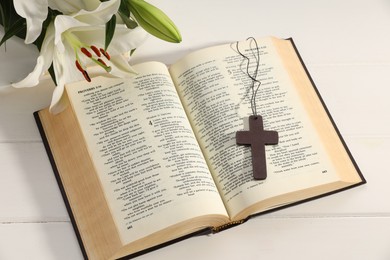 Cross, Bible and beautiful lily flowers on white wooden table, above view. Religion of Christianity