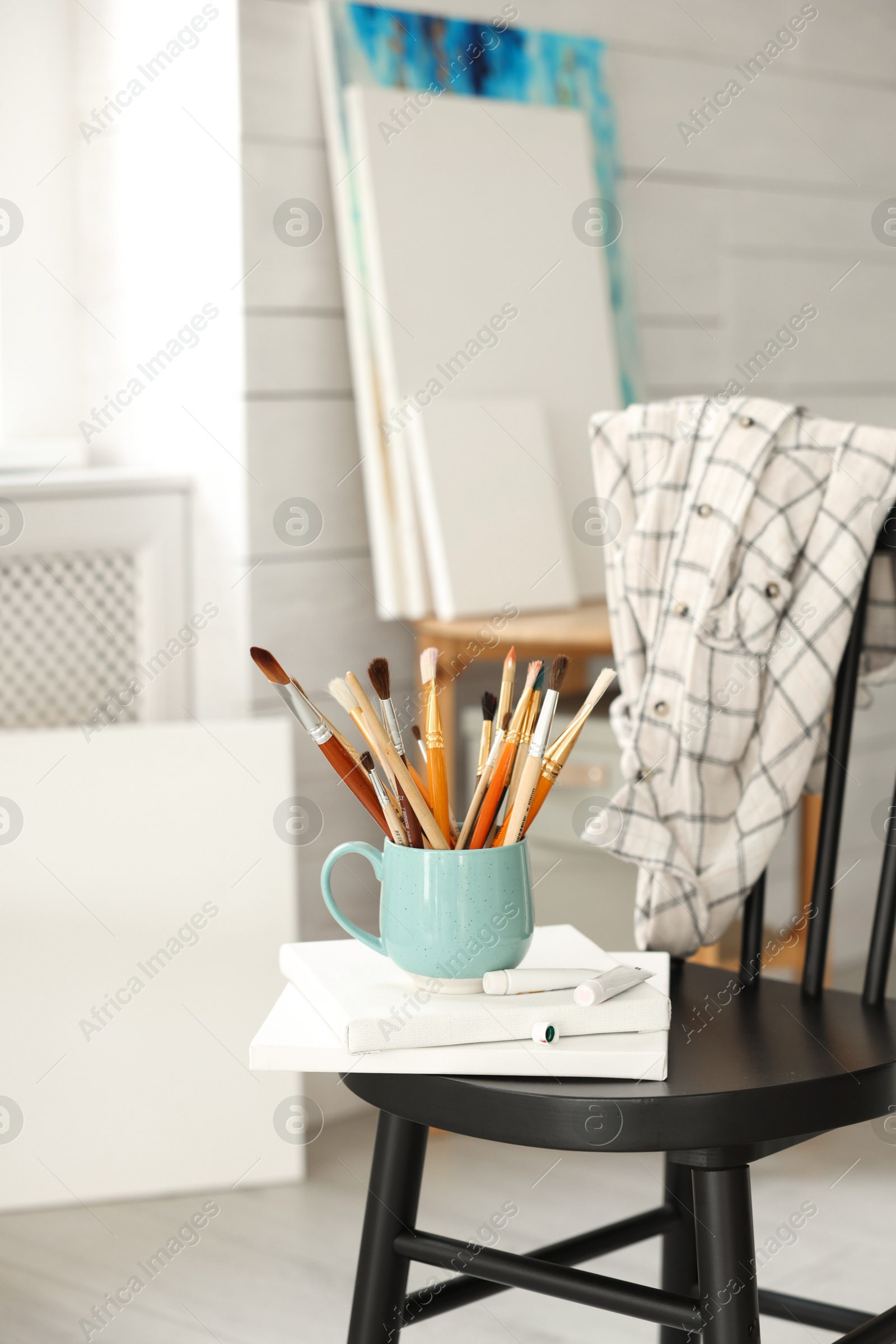 Photo of Canvases, paints and cup of brushes on chair in workshop