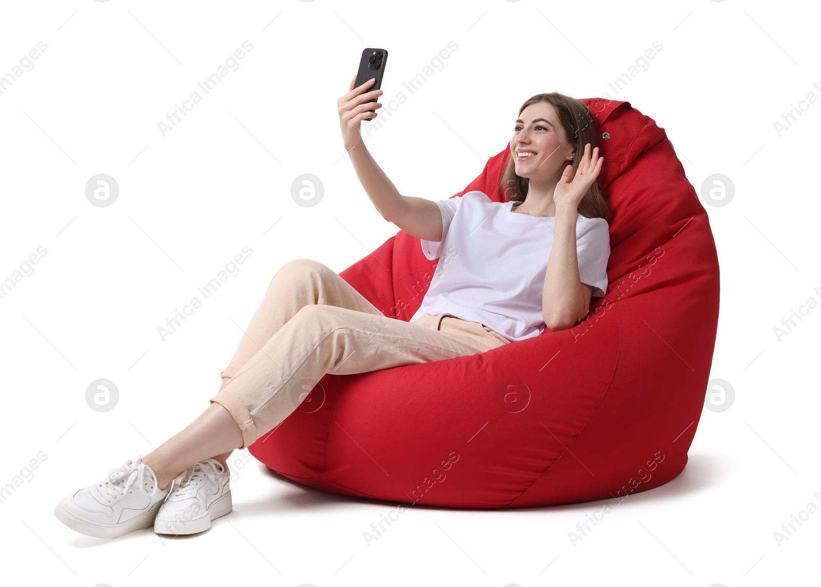 Photo of Smiling woman with smartphone having online meeting while sitting on red bean bag chair against white background